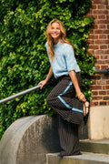 Woman in light blue sweater and striped pants standing against a brick wall with greenery.